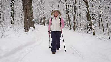 Energetic woman in warm white waistcoat with hood and black trousers spending her leisure with active nordic walking