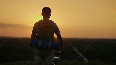 A man with a backpack and a bicycle. It stands and looks at the horizon where the sun sets. Cycling and adventure