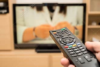 Watching TV and using black modern remote controller. Hand holding TV remote control with a television in the background