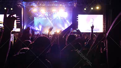 Many arms raised of fans in bright lights of stage, crowd of people waving arms and hold phone with digital displays