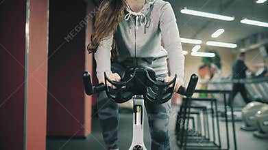 Young beautiful girl in gym, shaking her legs on cycling simulator, smiling at camera. The concept: to love sports, to