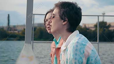 Two girlfriends relaxing by the pond sitting on the edge of a wooden pier and talking to each other. Joyful summer day