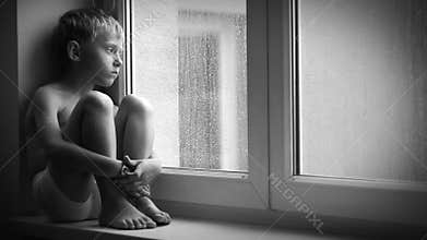 Black and white footage of a sad boy sitting on the windowsill during downpour, unable to get out of the apartment.