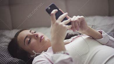 Young woman lies on a bed with a smartphone in hands, earphones plugged-in, girl listens to the music or podcast.