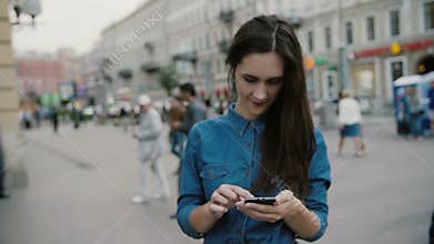 Modern technology. Beautiful brunette young woman in a denim dress using her smartphone in a busy street. Slow mo