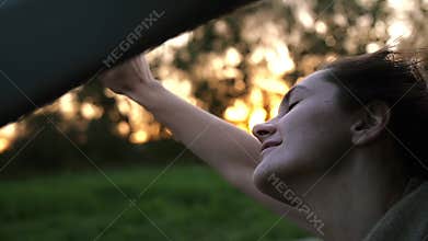 Pretty woman in a car, closing her eyes in happiness, hand playing in the air. Sun sets. Wind blows hair. Slow mo
