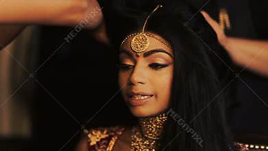 Alluring Indian bride holds her delicate fingers behind her temples while women get her hair ready