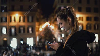 Young attractive woman standing in the city centre at evening and use the smartphone. Crowd and lights on the background