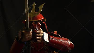 A samurai in red armor, a helmet and a defensive mask in the form of a demon catches the palm of the katana and selects