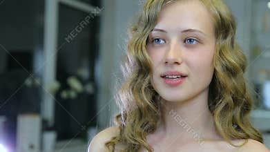 Beautiful young girl with long curly blond hair and blue eyes looks herself before makeup