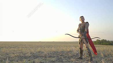 A brave archer in armor and a red cloak is standing with a stretched bow and looking at the camera, slow motion
