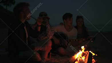 Picnic of young people with bonfire on the beach in the evening. Cheerful friends singing songs and playing guitar