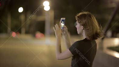 Beautiful young woman taking picture on smartphone at night street