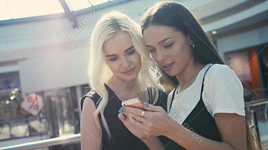Two beautiful young female shopaholics surfing the internet in search of discounts in shopping mall, young student girls