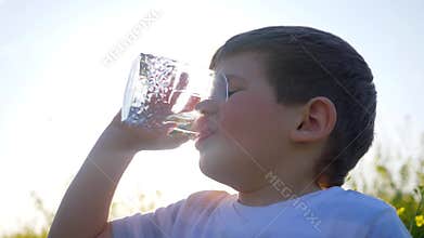 Happy small guy drink clean water on nature on background field flowers, little boy drinking from glass outdoors,child