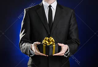 Theme holidays and gifts: a man in a black suit holds exclusive gift wrapped in a black box with gold ribbon and bow on a dark