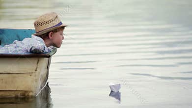 Boy wearing wicker hat sitting in the boat setting his paper boat on a journey