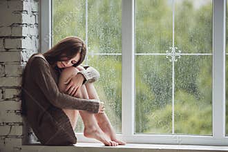 Beautiful young woman sitting alone close to window with rain drops. and sad girl. Concept of loneliness