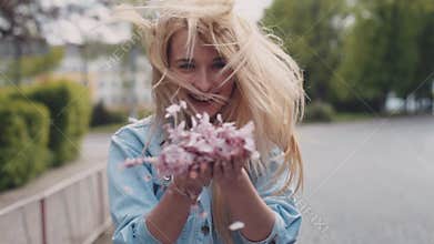 European young blonde attractive girl is moving down the park, then she turns and throws the petals of cherries blossom