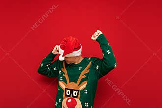 Cheerful young man in santa hat and green sweater dancing on red background. Cheerful guy having fun at Christmas party. Isolated