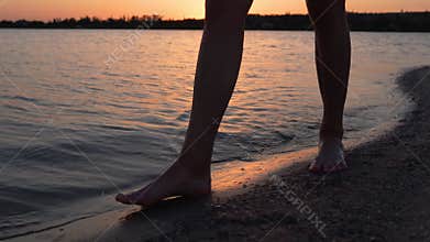 Male legs and feet walking along lake or pond on sandy beach in the dusk. Men move on shore alone. Calm water on
