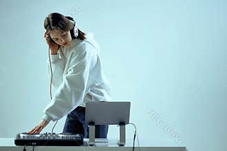 Cool young girl DJ mixes music on a mixing console and laptop, in stylish clothes, on white blue background.