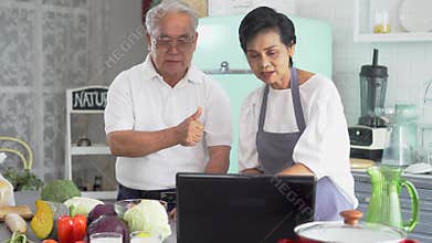 Senior Asian couple using a laptop while cooking in kitchen at home. 70s Elderly man and woman in relationship
