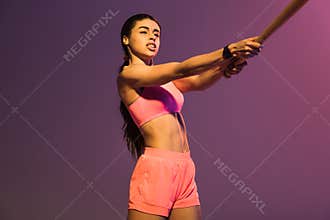 Sportive african american girl in pink sports bra playing baseball on purple background