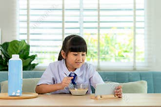 Asian elementary school student girl in uniform eating breakfast cereals with milk and looking cartoon in smartphone to