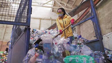 Footage of a young woman in yellow jacket and gloves scoops using shovel used plastic bottles at recycling factory. Huge