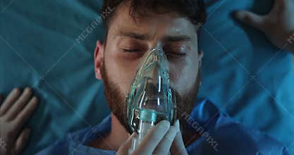 Close up of medics hands putting on ill bearded man oxygen inhalation mask. Top view of unconscious guy in 30s mumbling