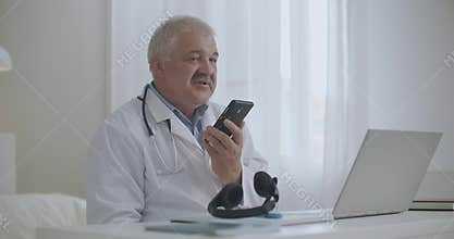 Male doctor is talking by speakerphone in mobile phone in his office in clinic, recording voice message, consulting