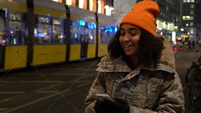 Young woman teenager using her cell phone at night with trams by Alexanderplatz Station, Berlin, Germany