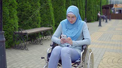 Young attractive muslim woman in a traditional scarf disabled in a wheelchair uses a smartphone sitting in the park