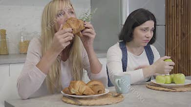 Portrait of upset plump Caucasian woman looking jealously at slim blond friend eating tasty croissants and biting apple