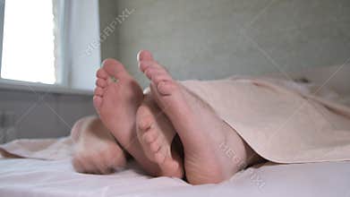 Close-up of the feet of a young couple sticking out from under the covers in the bedroom. Bare feet caress each other