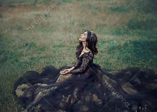 lady in black elegant dress sits on green grass. eyes closed, enjoys nature.