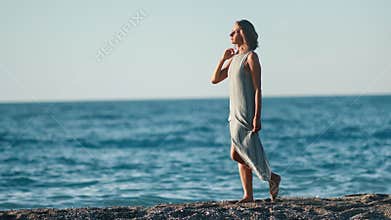 Attractive tanned travel woman wearing dress walking on beach at sunset full shot