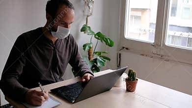 Young man wearing medical face mask in home office due to coronavirus spread, taking notes while working from computer in living