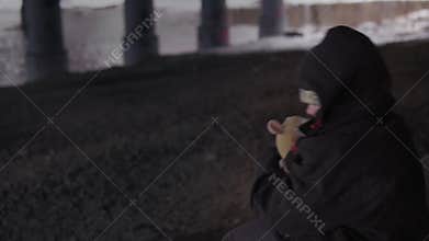 Helpless street child with toy sits in winter under bridge near the river.