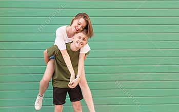 Smiling young man wearing casual clothing wears a happy girl on his back, looks at the camera and has fun. Stylish couple having