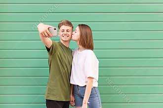 Loving young couple takes selfie on colored background, girl kisses boy, young man smiling and looking at the camera smartphone,