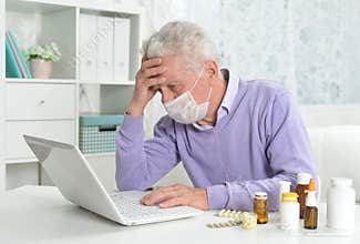 Sad sick senior man with facial mask using laptop