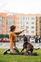 Stylish hipster girl dresses the dog on the lawn against the background of urban landscape, looks at the dog and smiles. Happy