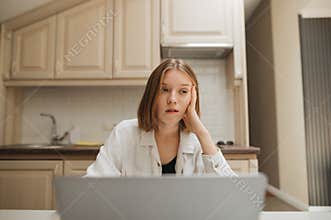 Attractive girl uses a laptop at the table at home and looks away with a unhappy face. Portrait of a bored girl working in the