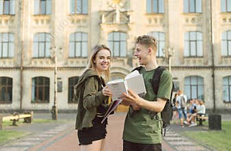 Happy couple standing on background of university building with books and exercise books in hand, guy shows book to girl, she is