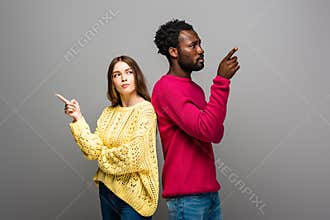 Interracial couple in knitted sweaters standing back to back and pointing with fingers on grey background