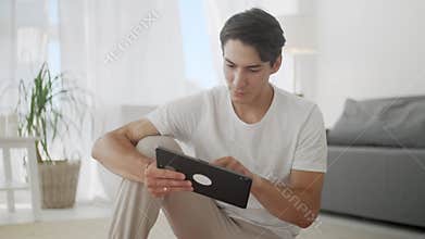 Smiling Young Man Surfing Internet on Tablet While Sitting on Exercise Mat During Relaxing After Workout at Home.