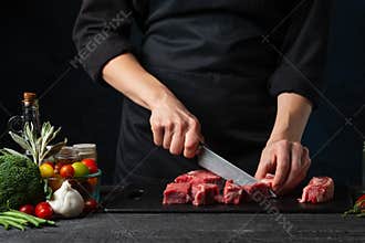 The professional chef cutting with knife fresh beef meat on the black board on dark blue background. Backstage of preparing rib-