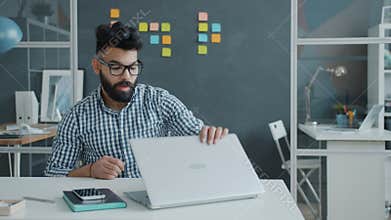 Unhappy Middle Eastern employee using laptop then hitting table with fist and leaving workplace